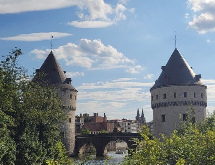 Das Bild zeigt die historischen Broelt&uuml;rme in Kortrijk, Belgien. Zwei runde, mittelalterliche T&uuml;rme mit kegelf&ouml;rmigen D&auml;chern stehen am Ufer eines Flusses und sind durch eine Steinbr&uuml;cke verbunden. Die Szene wird von B&auml;umen und B&uuml;schen im Vordergrund umrahmt, und der Himmel ist mit einigen Wolken bedeckt, was eine friedliche Atmosph&auml;re schafft.