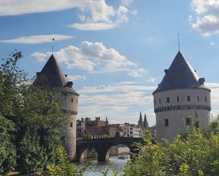 Das Bild zeigt die historischen Broelt&uuml;rme in Kortrijk, Belgien. Zwei runde, mittelalterliche T&uuml;rme mit kegelf&ouml;rmigen D&auml;chern stehen am Ufer eines Flusses und sind durch eine Steinbr&uuml;cke verbunden. Die Szene wird von B&auml;umen und B&uuml;schen im Vordergrund umrahmt, und der Himmel ist mit einigen Wolken bedeckt, was eine friedliche Atmosph&auml;re schafft.
