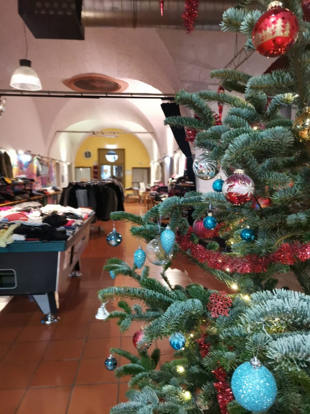 Weihnachtsbaum im Vordergrund. Im Hintergrund ein Raum mit vielen Kleiderstapeln auf Tischen