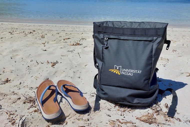 Rucksack der Universität Passau neben Flipflops am Strand 