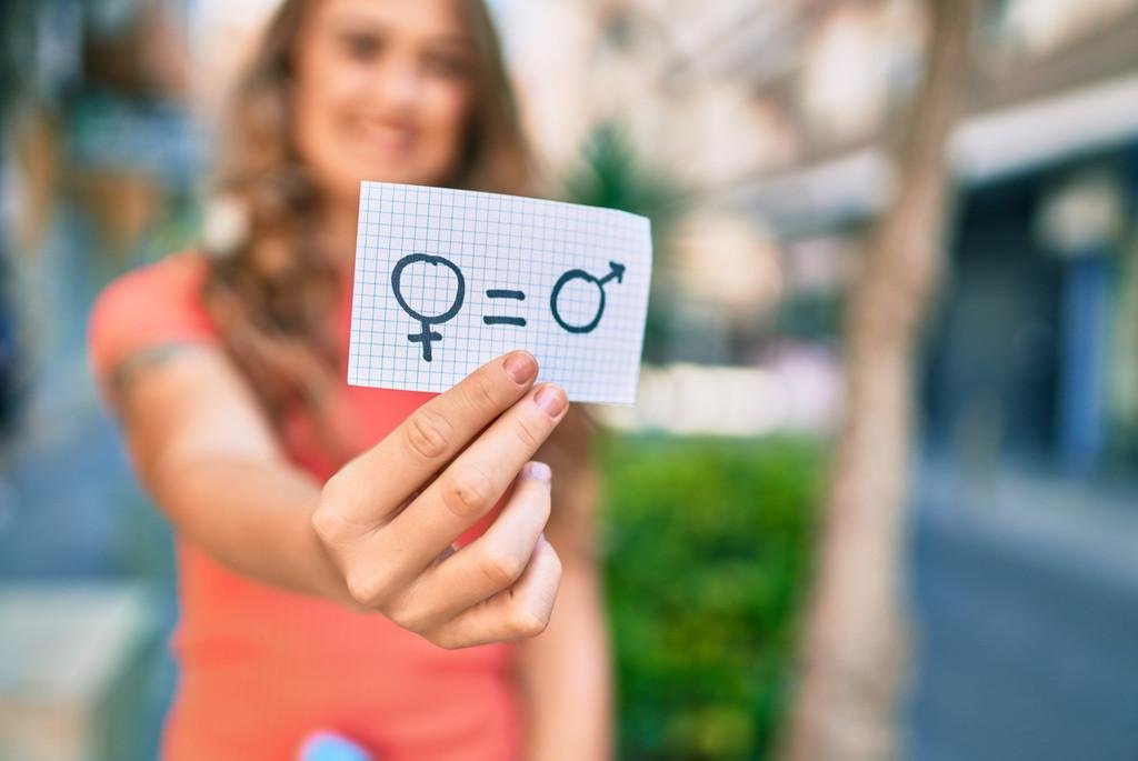 Symbolbild: Eine Frau, die man nur verschwommen im Hintergrund erkennen kann, hält einen Zettel mit einem Symbol für Geschlechtergleichheit in die Kamera.