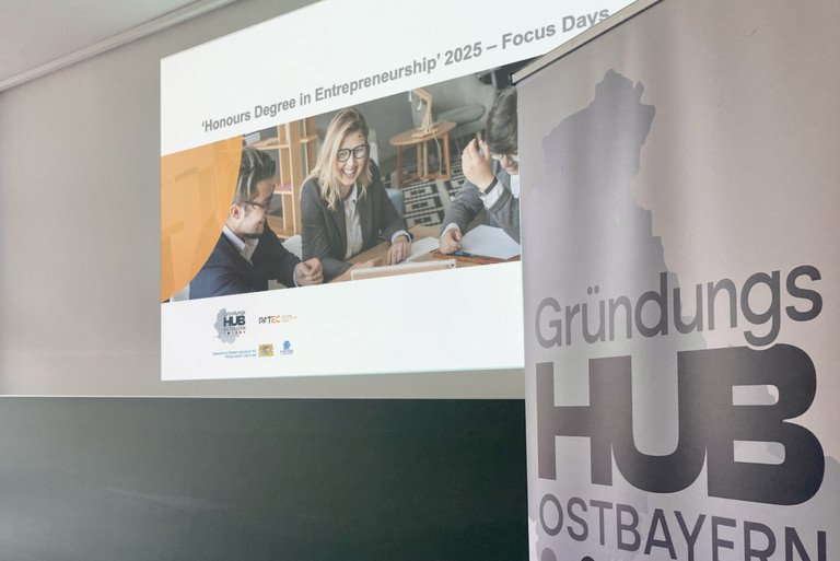 Im Vordergrund ist ein Roll-up zu sehen, auf dem das Logo des Gründungshubs Ostbayern gedruckt ist. Im Hintergrund ist eine Powerpoint-Folie mit einem Bild und der Aufschrift "Honours Degree in Entrepreneurship" und "Focus Days" abgebildet.