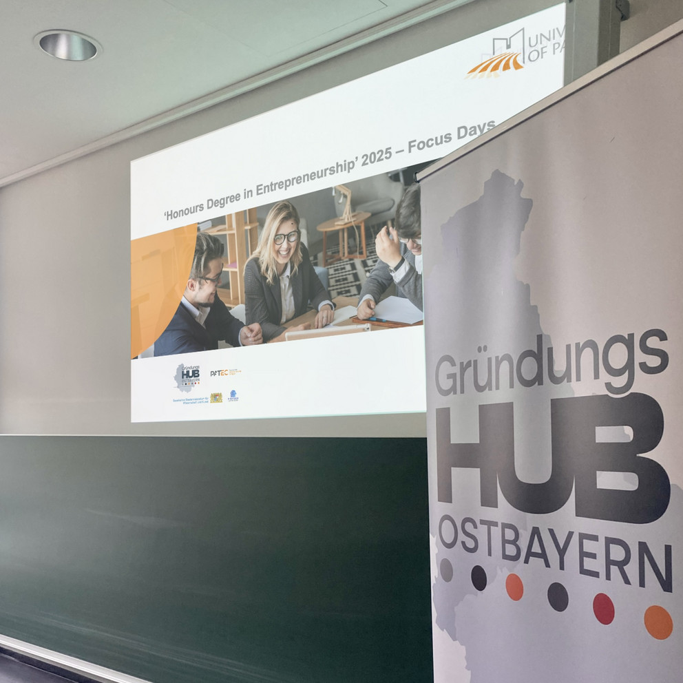 Im Vordergrund ist ein Roll-up zu sehen, auf dem das Logo des Gründungshubs Ostbayern gedruckt ist. Im Hintergrund ist eine Powerpoint-Folie mit einem Bild und der Aufschrift "Honours Degree in Entrepreneurship" und "Focus Days" abgebildet.