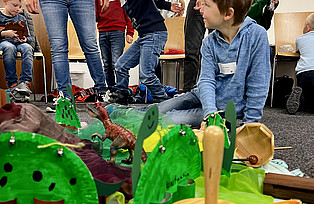 Kinder sitzen vor einer Landschaft mit Dinosauriern und kleinen Instrumenten auf dem Boden.