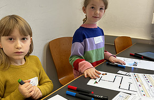 Zwei Kinder sitzen an einem Tisch. Auf dem Tisch liegen Blätter mit Wegen aus schwarzen Linien für die Roboter.
