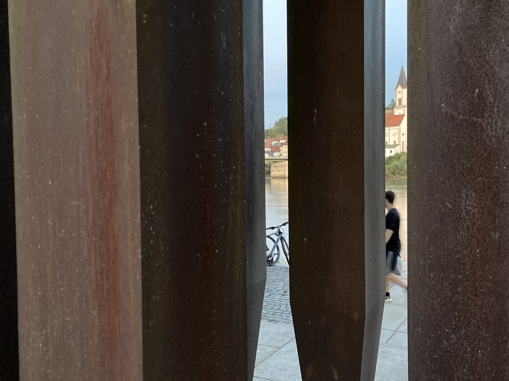 Nahansicht durch die Skulptur hindurch, im Hintergrund jenseits des Inns zu sehen ist die Kirche St. Gertraud