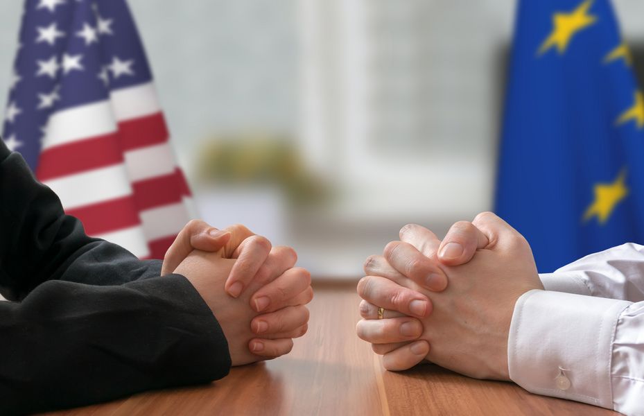 Symbolbild: Nahaufnahme von zwei Paar Händen mit amerikanischer und Europa-Flagge im Hintergrund